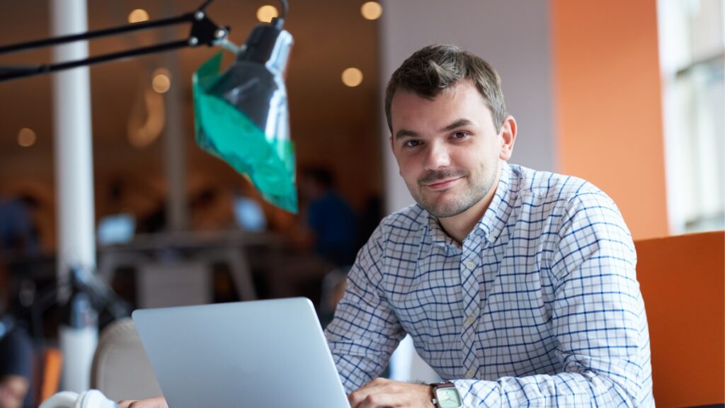 A man smiles at the camera while he works on his laptop
