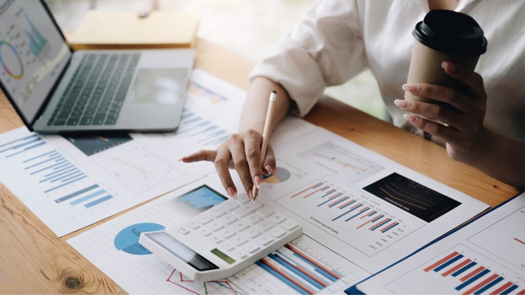 Someone usess a calculator with one hand and holds coffee in the other hand, on a table full of charts