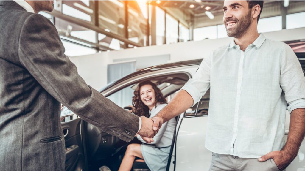 A man in a suit shakes the hand of another man in front of a woman in a car smiling at them