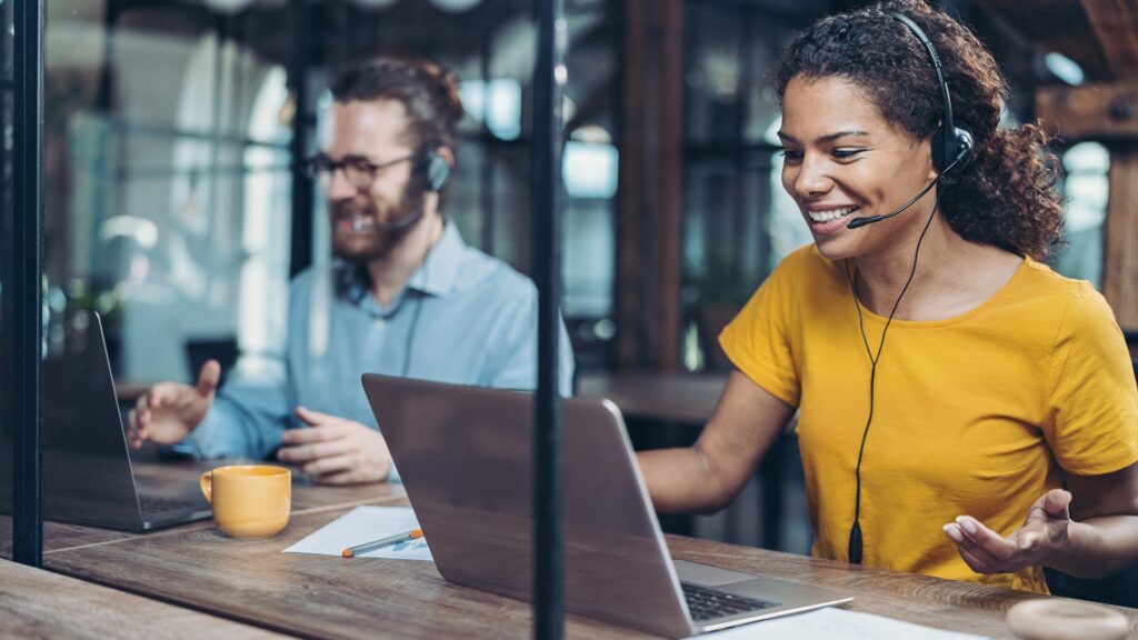 A man and a woman wearing headsets smile at their laptops