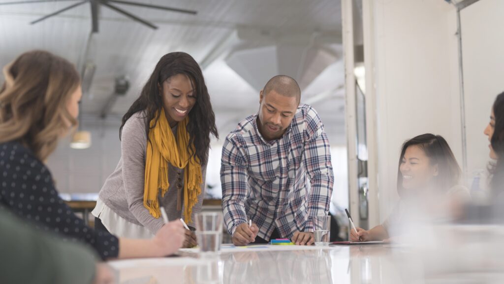 Two people standing, bending to write on the table, while others sits around the table