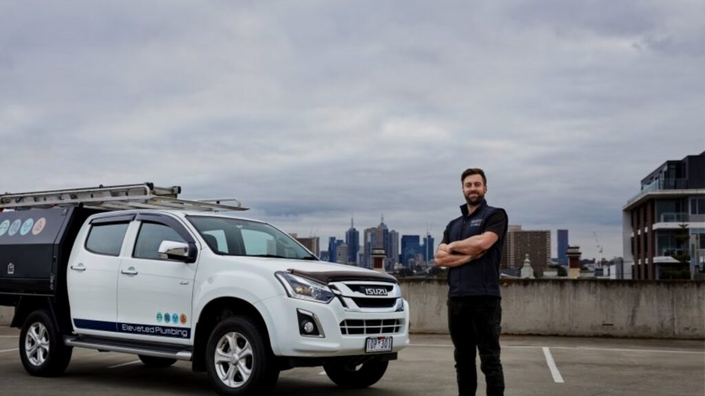 Owner of Elevated Plumbing standing confidently beside his branded company vehicle on a rooftop with a city skyline in the background