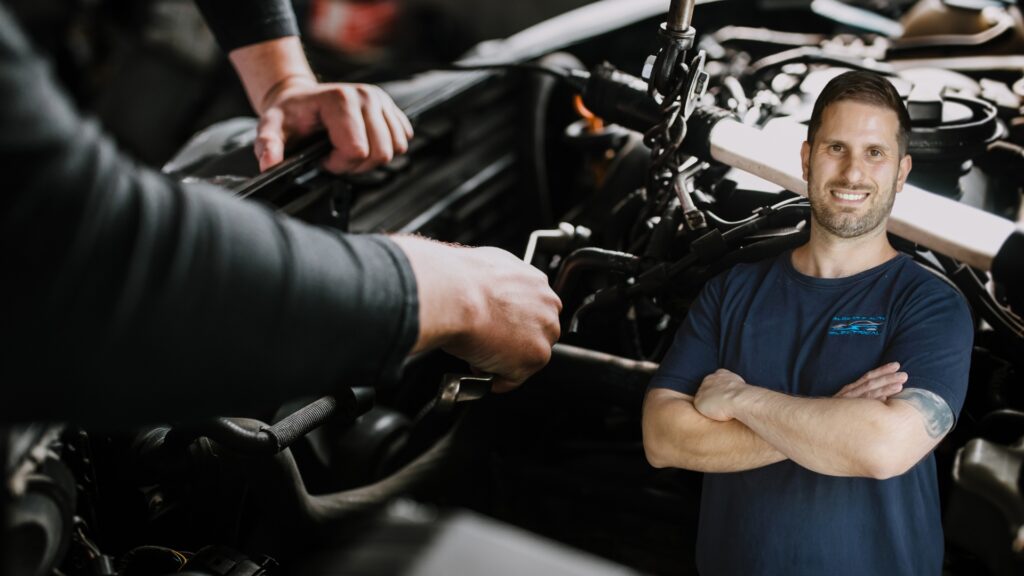 Business owner from Albert’s Auto & Electrical smiling confidently with arms crossed in front of a car engine