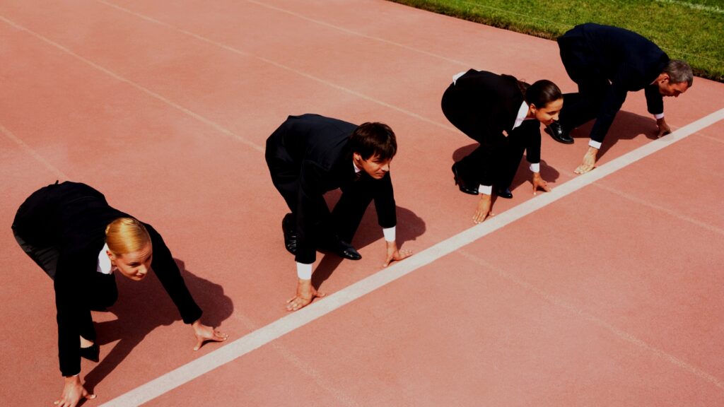 Four business professionals in suits crouch at a starting line on a track, ready to race