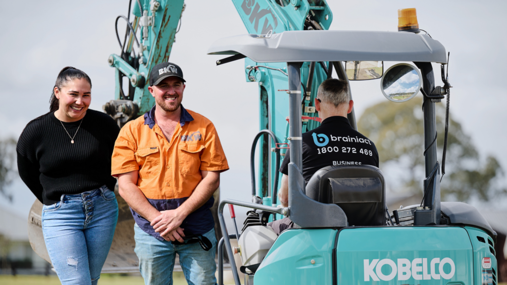 a Brainiact coach in a Kobelco excavator and two BKW Earthworks employees smiling and engaged