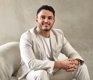 A man in a suit sits a on white couch, smiling