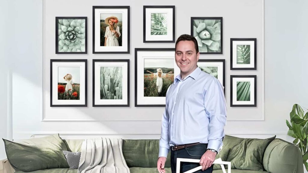 A man smiles in the foreground with picture frames on the wall and a couch in the background
