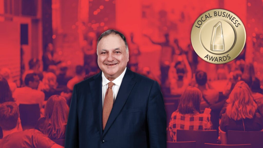 A man in a suit stands smiling in front of a background featuring an audience and a "Local Business Awards" logo. The image has a red color overlay.