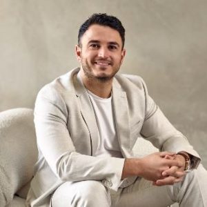 A man in a suit sits a on white couch, smiling